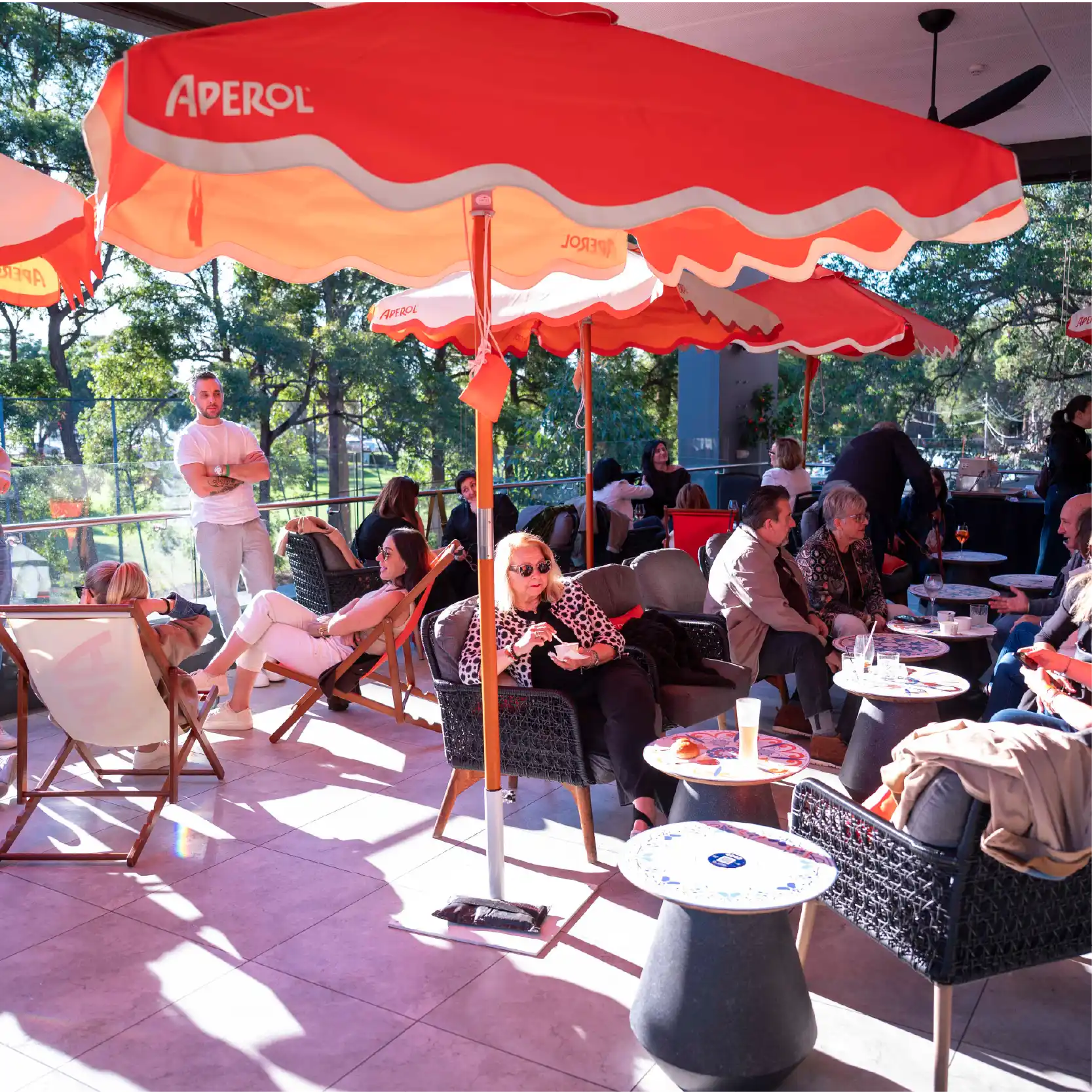 Sun shining through the trees, orange aperol umbrellas, people having food.
